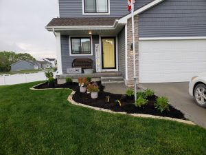 New dark mulch garden bed with limsetone edging and new plants lining the upper portion of a driveway on the left side of the house and around the sidealk and around the corner of the house. The home has a white garage door and is a two story grey sided home - Norwalk Seasonal - Des Moines, Iowa