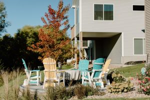 A nice circular stone laid patio about 15ft from the house with 3 blue and white wooden chairs and two yellow around a covered firepit. The patio is surrounded by a nice rock garden with flowers and plants as well as a walkway.