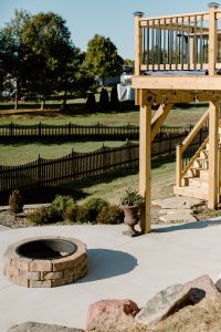 A concretee patio with a stone firepit all surrounded by a rock garden with large stones and plants. Also pictured is a second level deck with a staircase leading up to it and the fence of the neighboring yard. Poured Concrete.