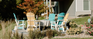 A nice circular stone laid patio about 15ft from the house with 3 blue and white wooden chairs and two yellow around a covered firepit. The patio is surrounded by a nice rock garden with flowers and plants as well as a walkway.