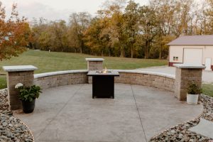 Textured concrete patio with a retaining wall providing seating and columns to be used as tables with a mobile gas firepit in the center. The patios is surrounded by rock and the patio has a lighted border