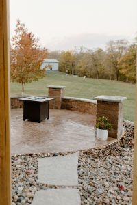 Large flagstone steps out to a patio with a retaining wall that is also used for seating. Also pictured is a gas firepit.