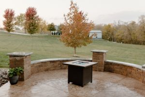 Gas firepit on a stamped and colored concrete patio with a retaining wall that doubles as seating. Also pictured is a gas firepit in the center of the patio and trees behind it.