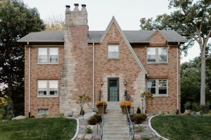 Large 2 sotry brick house with a tall chimney and staircase leading up to the front door with a nicely landscaped front yard.