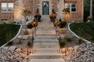 Staircase that is leading up to a dark gray decorative door on a brick house with a nice rock garden filled with large stones and plants. Also pictured are porch pots. Landscaping done by Norwalk Seasonal Services