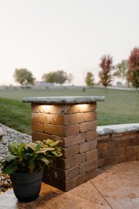 Closeup of a column of a brick retaining wall with a potted plant next to it and a light under the top stone on the column.